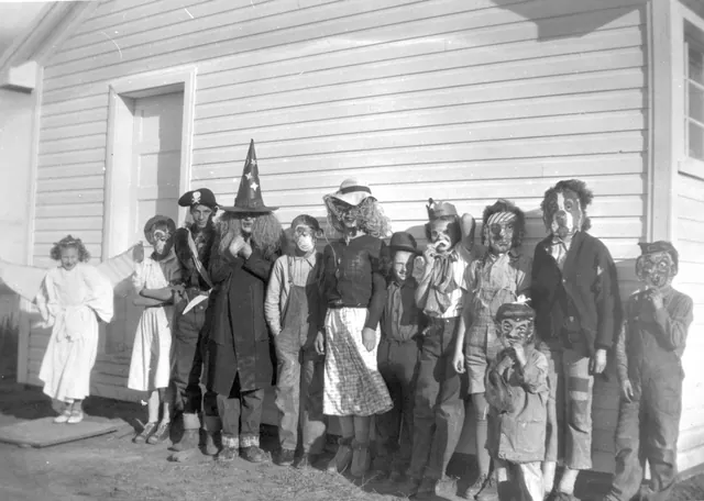 Children in Halloween masks and a witch hat lined up against a wooden wall around 1950